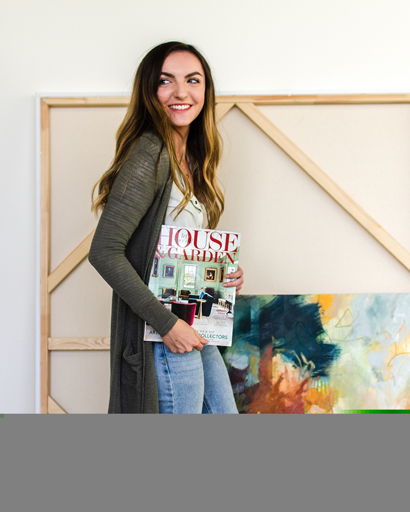 A young woman stands holding an issue of House And Garden magazine under one arm, posing in front of a larger canvas while wearing a long green cardigan. She has dark brown wavy hair and a wide smile.