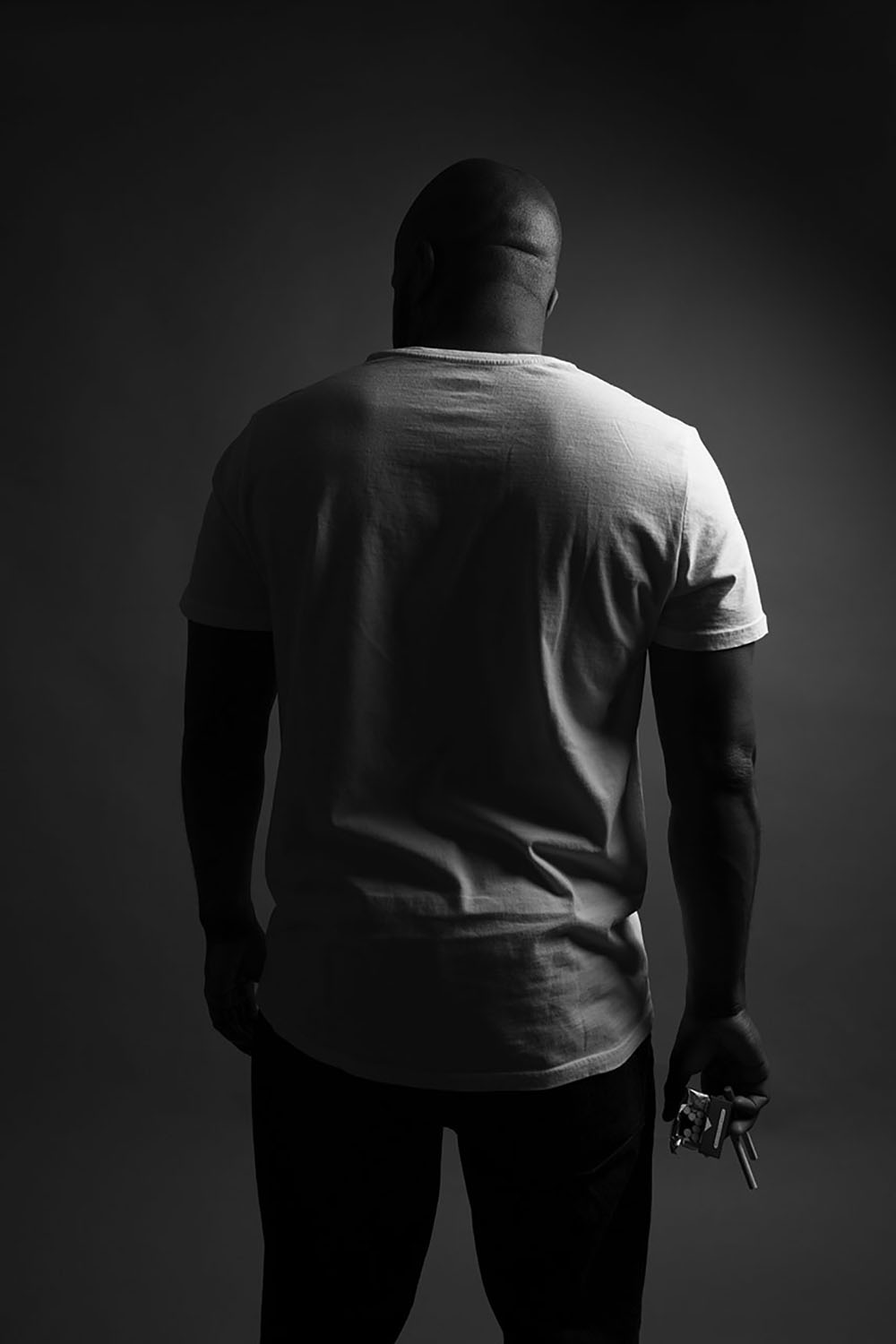 Image shows a black and white photograph of a Black man standing with his back to the camera, holding a pack of cigarettes in one hand.