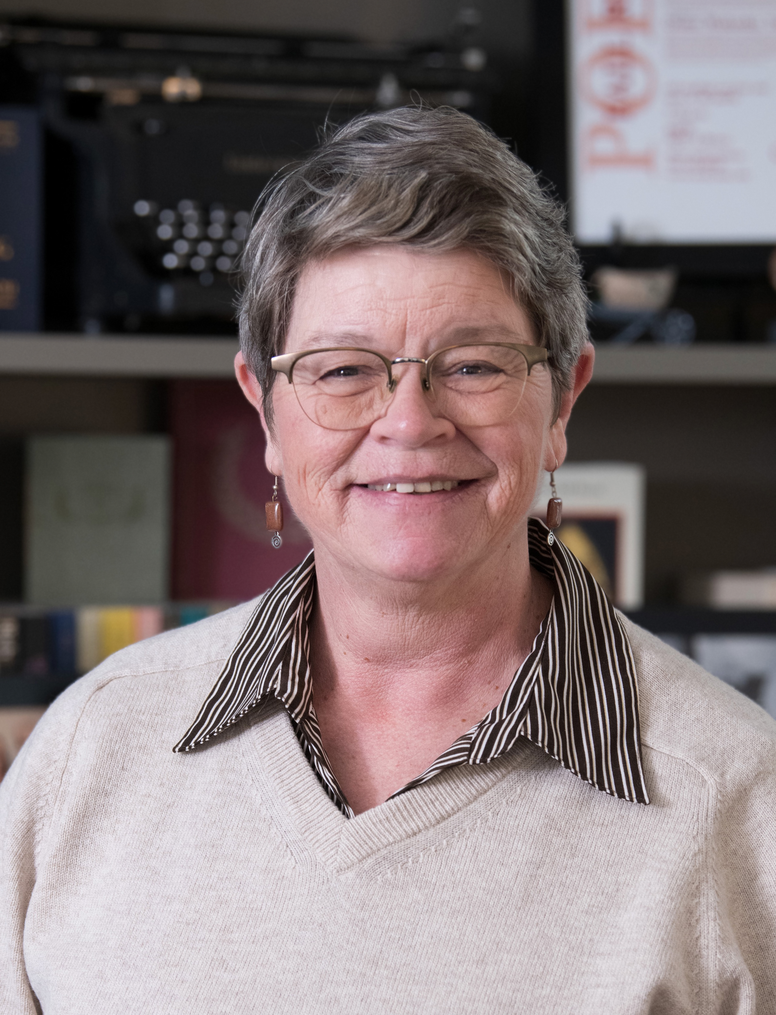 A woman with gray short hair, wearing brown glasses and a beige sweater, stands in front of a bookshelf, smiling at the camera