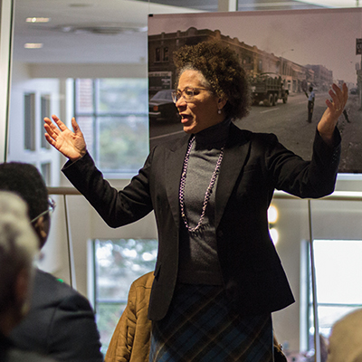 Tama Hamilton-Wray, a woman of color and professor, stands in a suit speaking in front of an audience in the LookOu! Art Gallery