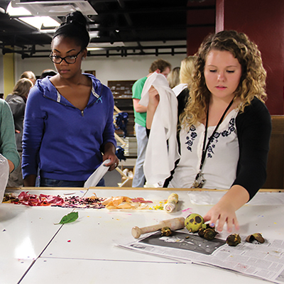 Two students stand in a studio space, working at a table to dye fabric.