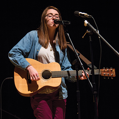 A young white woman in red pants and jean jacket stands on stage, holding a guitar and singing.