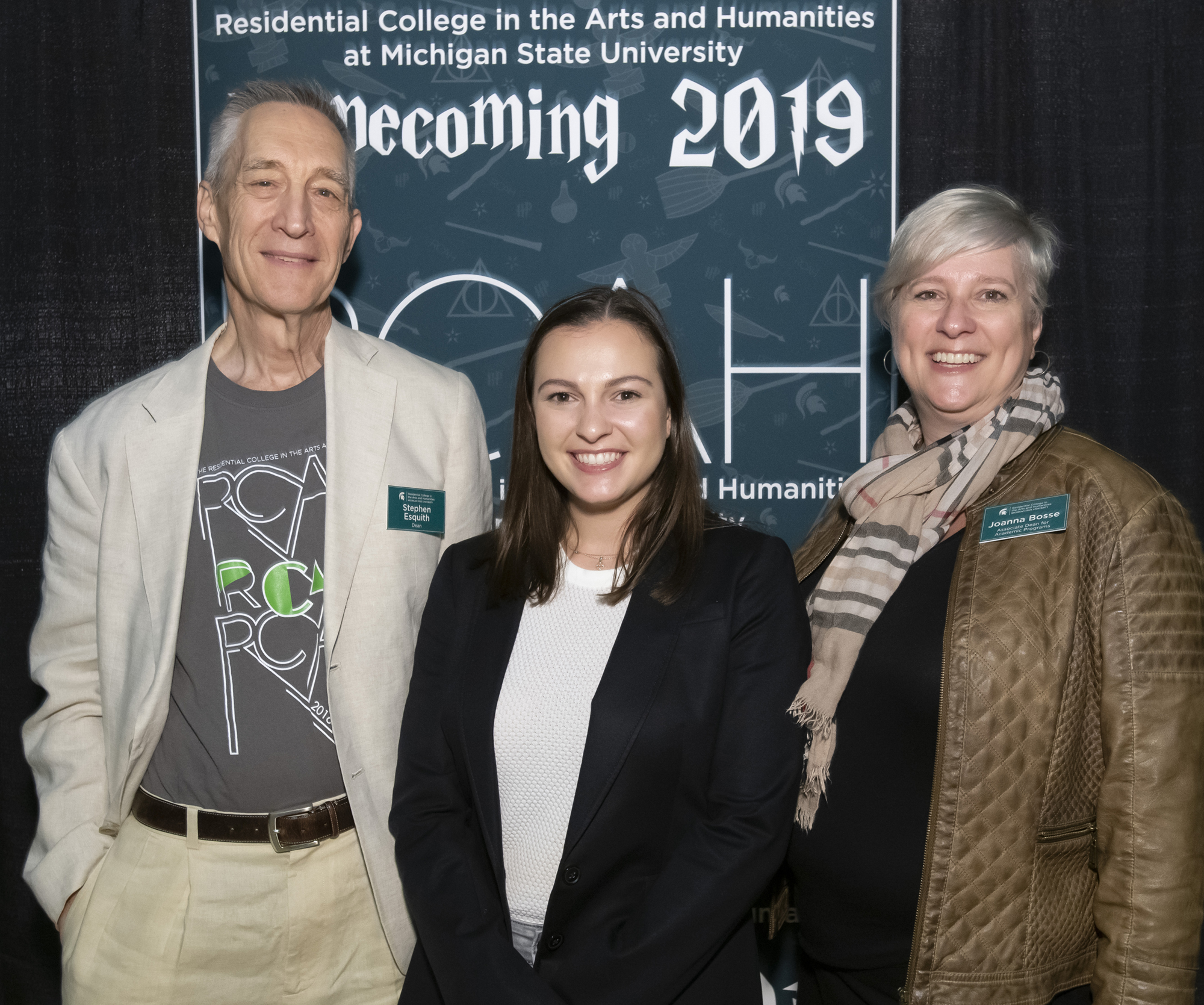 Three people stand in front of a banner: an older man in a pale suit, a young woman in a black suit jacket, and a white woman with short hair and a scarf.