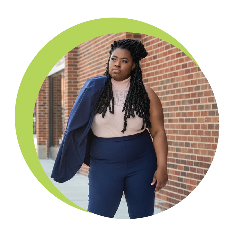 A young black woman in blue pants with matching blazer tossed over one shoulder stands proud, a brick wall and urban background visible.