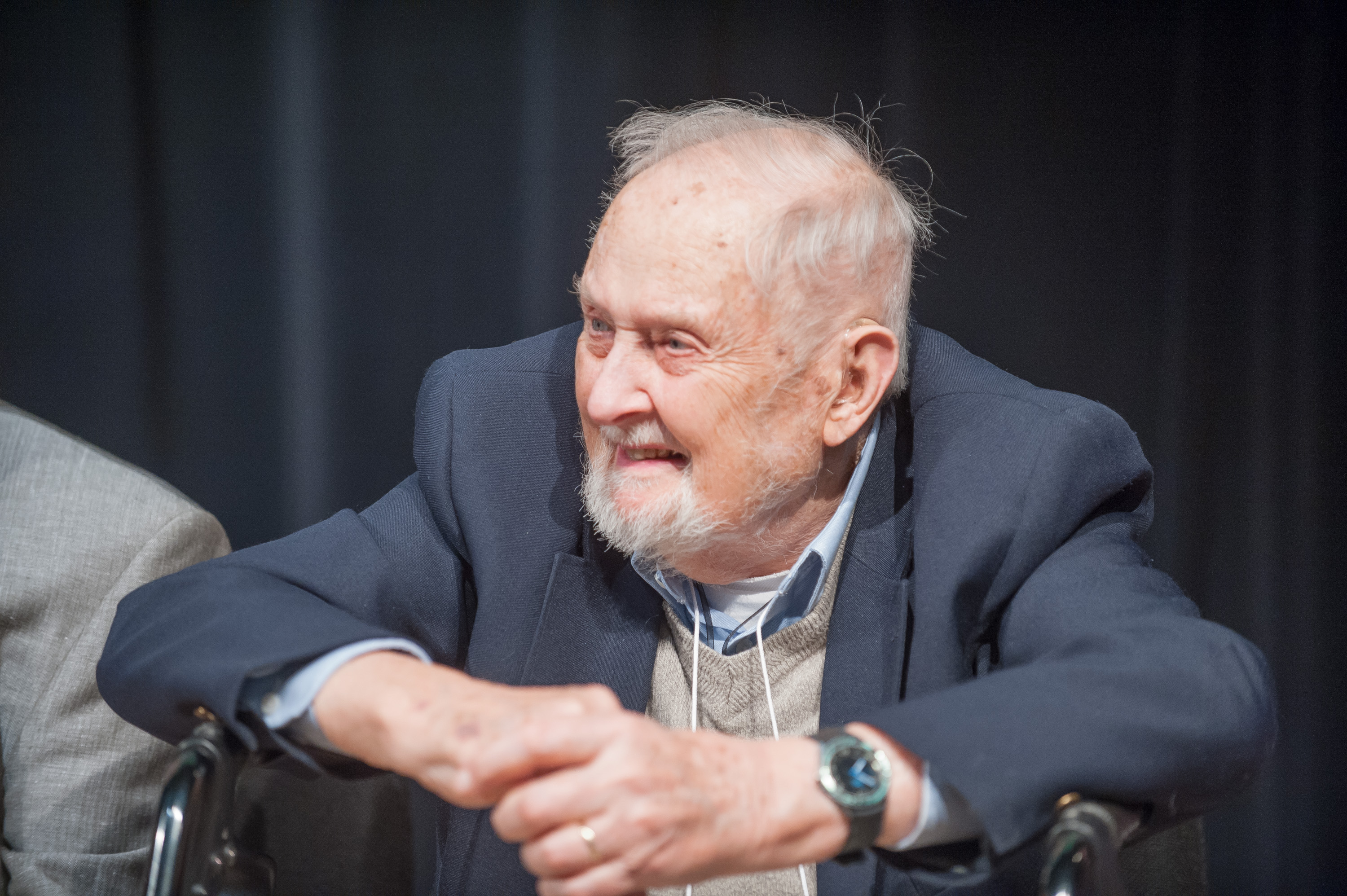 An elderly man dressed in a blue suit and seated on a stage with his arms resting on his walker smiles at the audience.